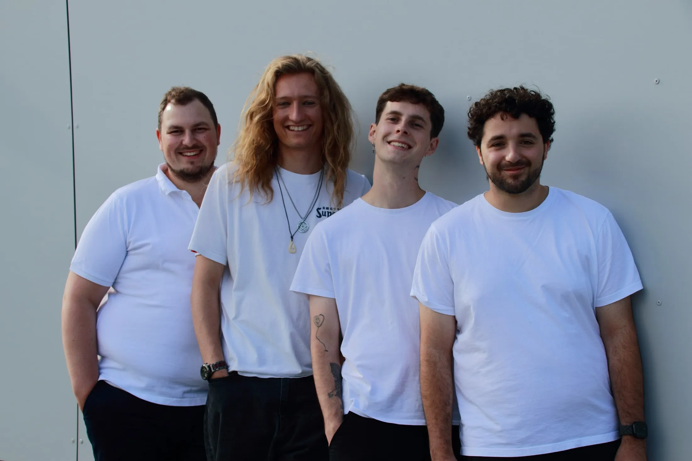 Die vier Baltigos stehen lächelnd in weißen Shirts vor einer hellen Wand